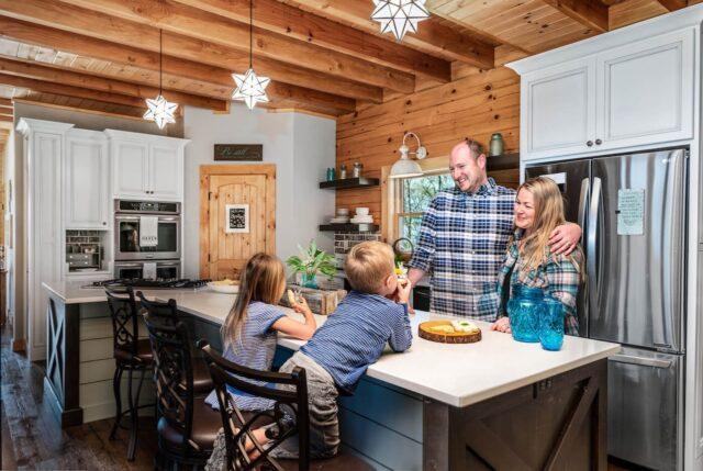 Photo of family in open floor plan kitchen with large island exposed wood ceilings with log support beams. The exterior wall of the kitchen is exposed log