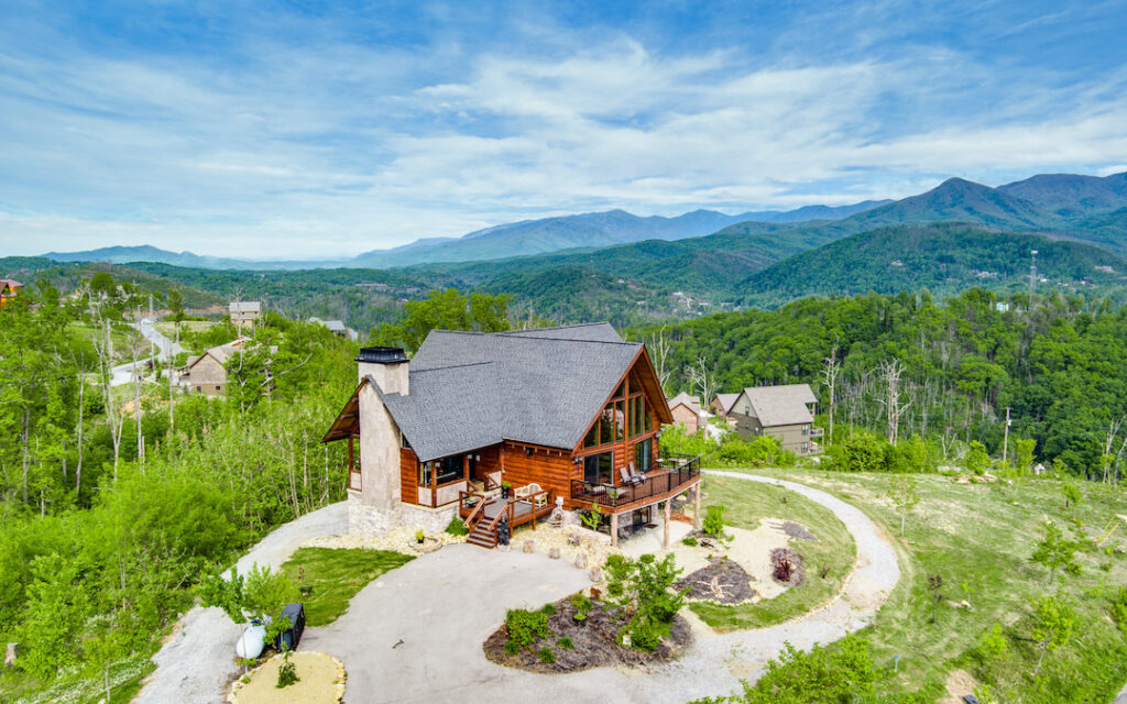 Aerial photo of log home on a hill. An exterior screened porch sports a stone fireplace and is the main entrance to the home. The home has a covered porch on the lower level and an upper open deck outside the great room of the home