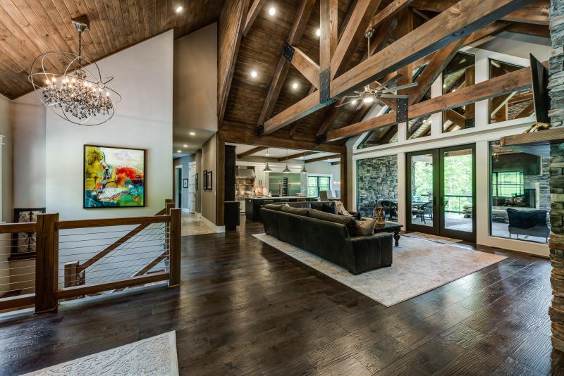 Photo of great room and kitchen of log home with exposed light wood ceiling and dark a-frame trusses. White framed glass windows lead to an porch