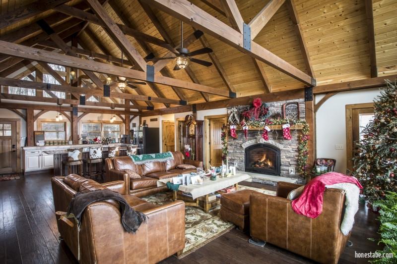 Photo of great room and kitchen of log home with exposed light wood ceiling and dark a-frame trusses. White framed glass windows lead to an porch