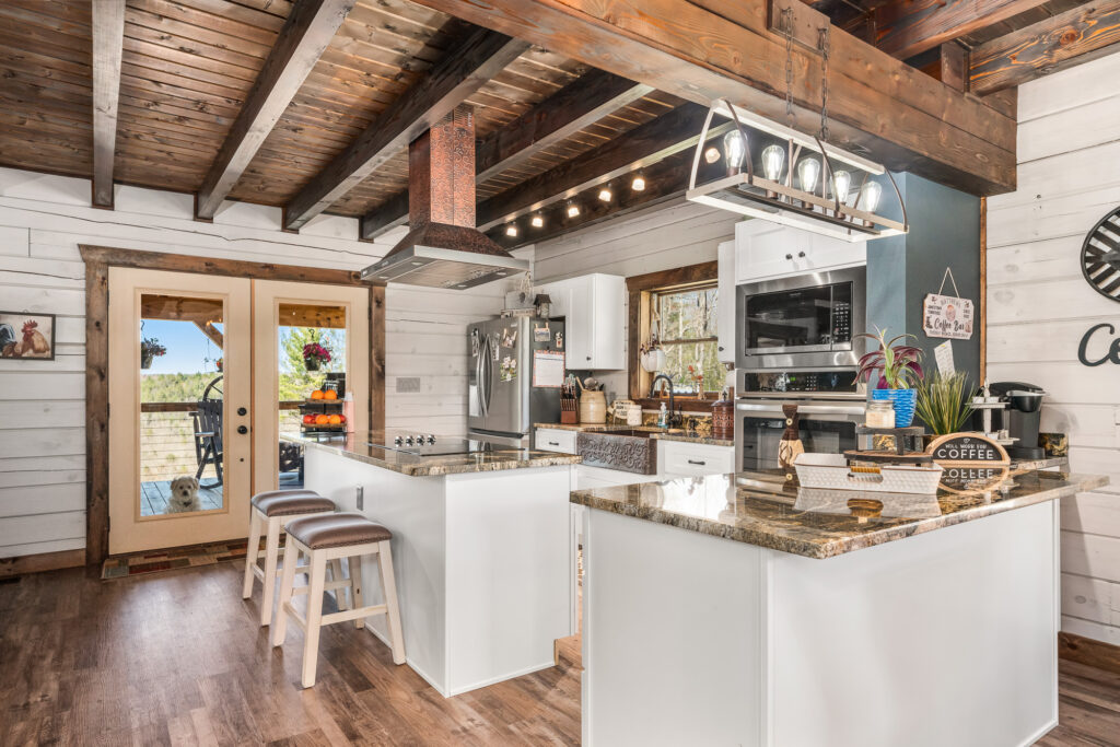 Photo of open kitchen set under a loft area of a log home. The interior walls are exposed wood painted a bright white. The ceiling and ceiling supports are dark heavy wood