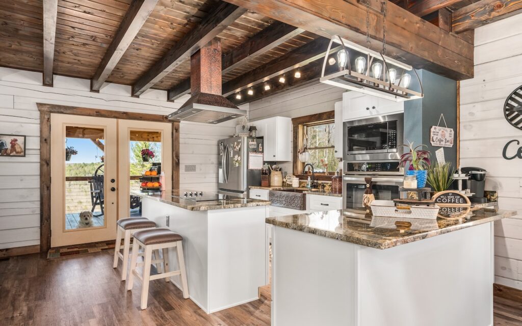 Photo of open kitchen plan with whitewashed wood walls, dark hardwood floors and dark hardwood support beams and ceiling. The main support beams are large square cut logs. Wood framed doors lead off the kitchen to an outdoor living space.