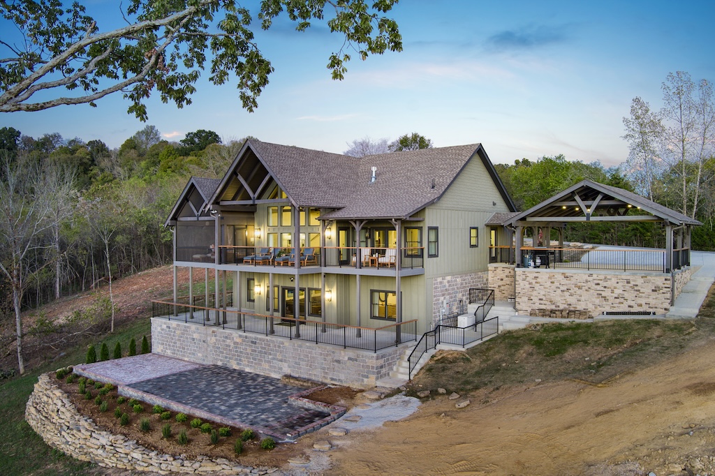 Photo of timber frame home with stone and wood exterior and three car garage bay attached to the home by an enclosed breezeway. Photo taken at sunset