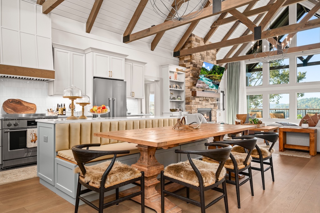 Photo of great room and kitchen of log home with exposed light wood ceiling and dark a-frame trusses. White framed glass windows lead to an porch