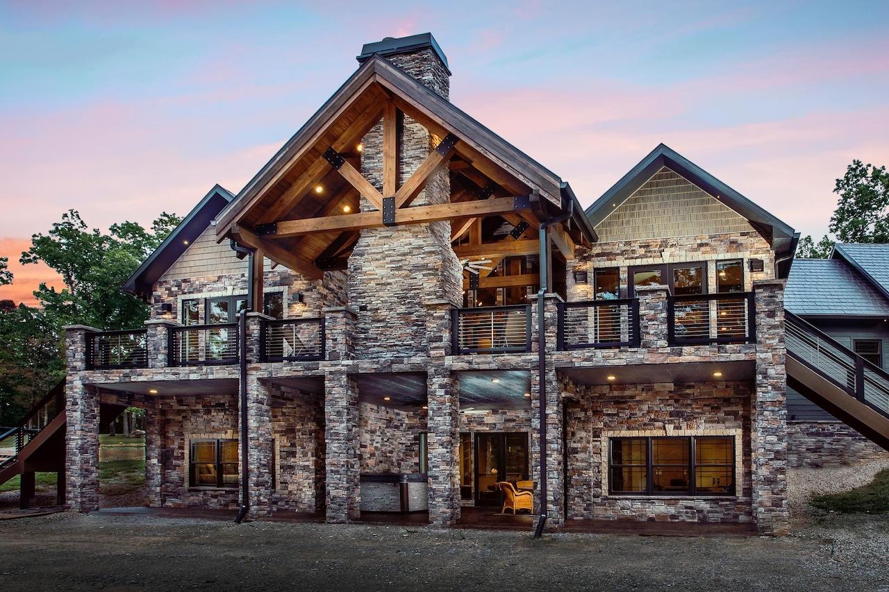 Photo of great room and kitchen of log home with exposed light wood ceiling and dark a-frame trusses. White framed glass windows lead to an porch